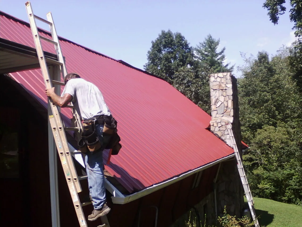 Roofer on a ladder beside a red metal roof and stone chimney — Soffit & Fascia Repair by Roofing Brothers in Bloomsbury